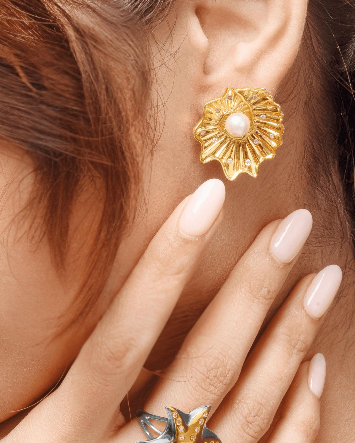 A close-up of a model wearing a textured gold flower-shaped stud earring with a white pearl center, with her hand resting near her face.