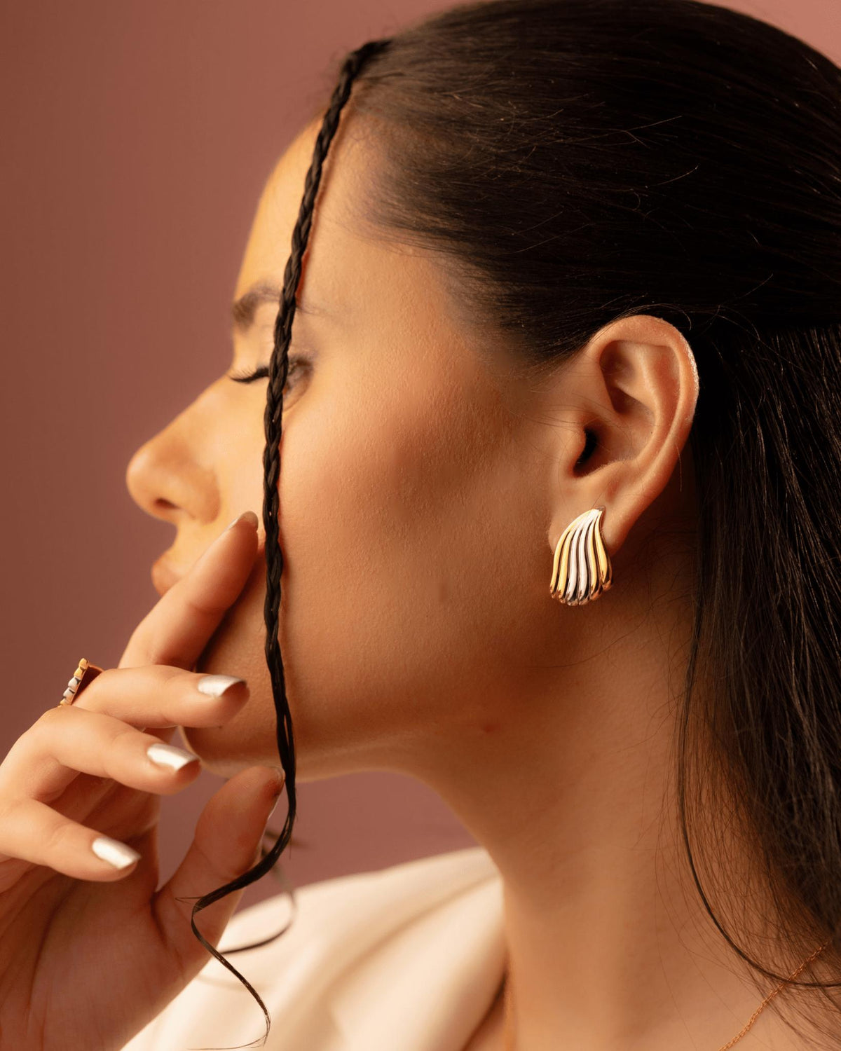 A close-up side profile of a woman wearing a gold wave-shaped stud earring and a gold ring, with her dark hair styled in a braid against a warm dusty rose background.