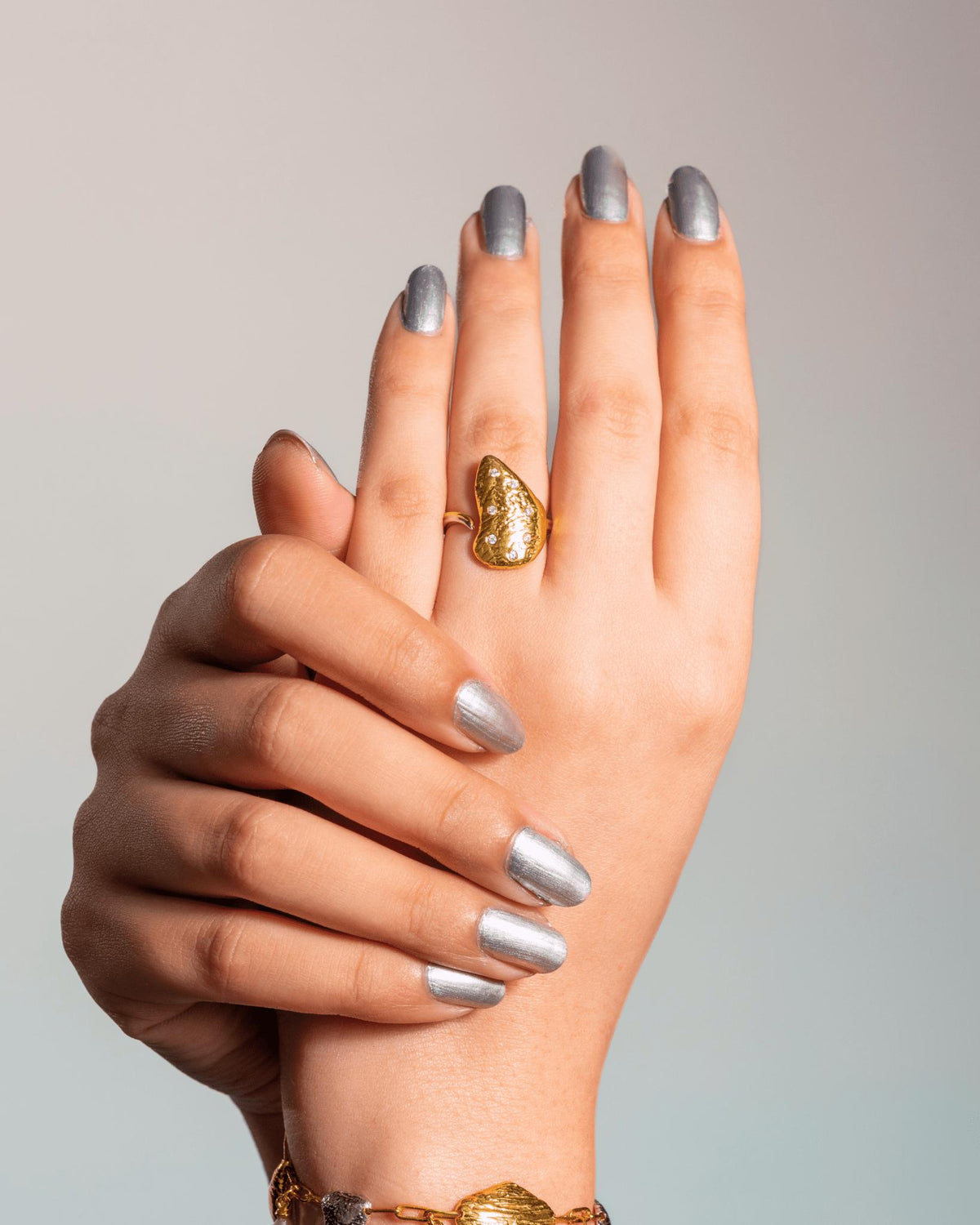 A close-up of a person's hands with silver manicured nails, wearing a textured gold ring, inspired by Oyster's Shell, on the middle finger, set against a neutral light gray background.