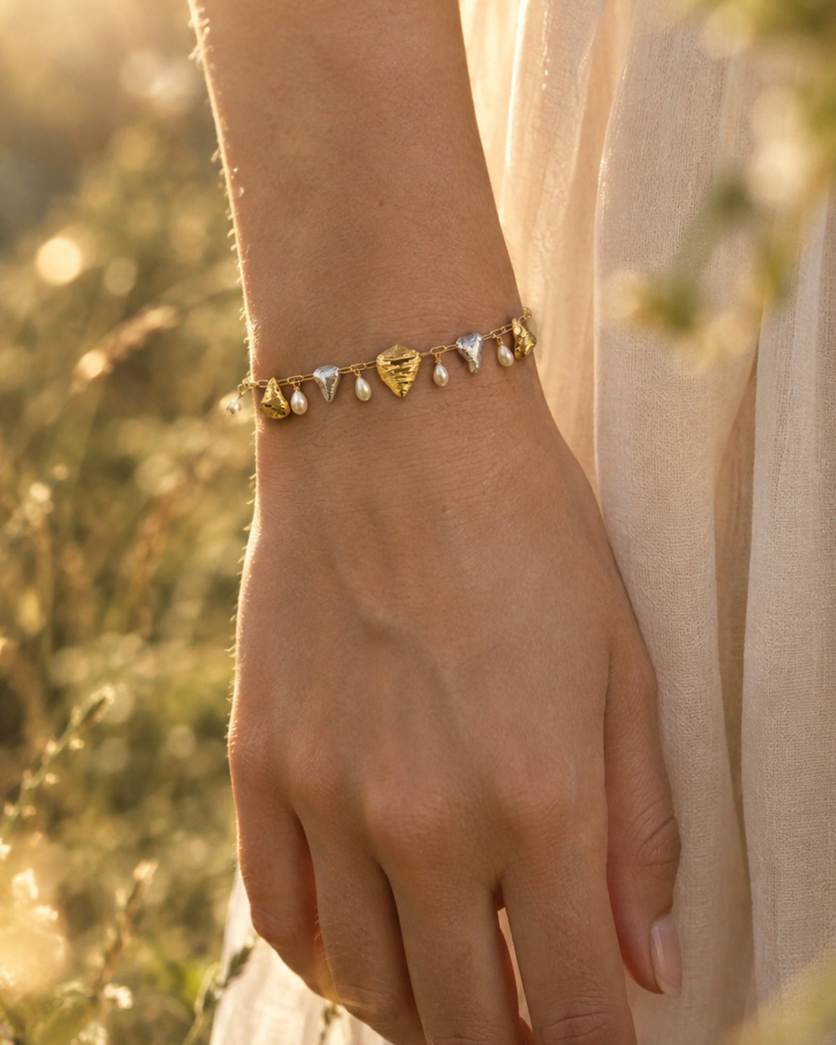 Model wearing dual-tone gold & silver oyster shell charm bracelet with pearls, outdoors in golden sunlight for brand story