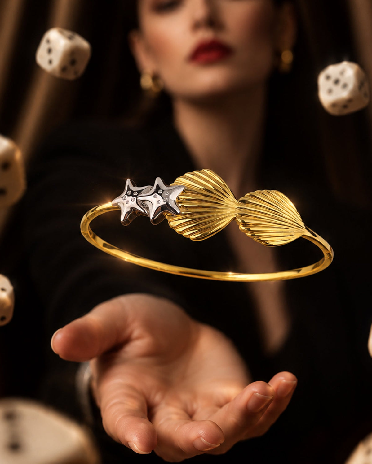 Floating dual-tone gold vermeil seashell & starfish cuff bangle above open hand with floating dice in dark mood setting
