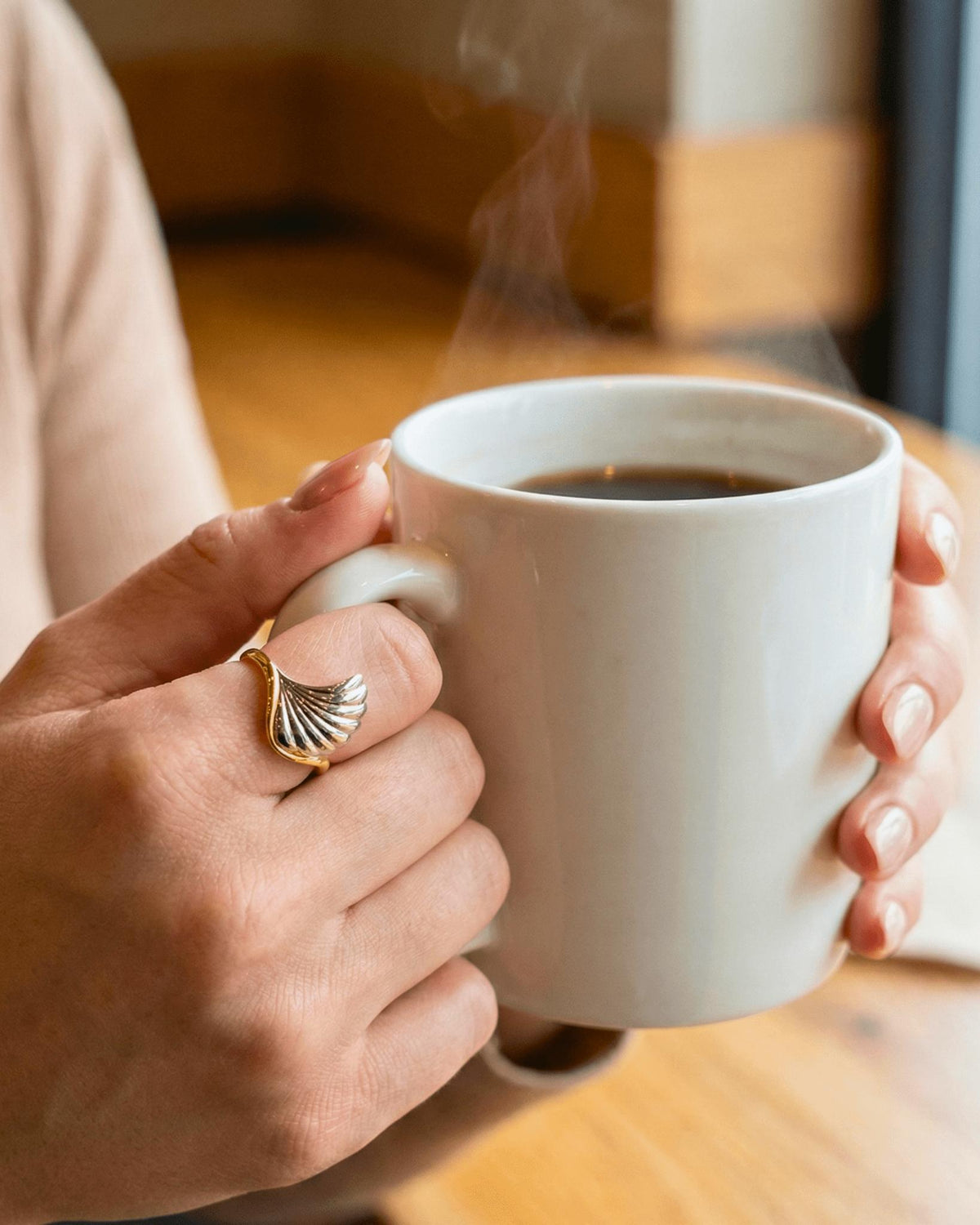 A close-up lifestyle shot of a person holding a steaming white ceramic mug with both hands, wearing a gold ring with a textured, wave flow design on the ring finger. The background is a softly blurred indoor setting with warm wooden tones.