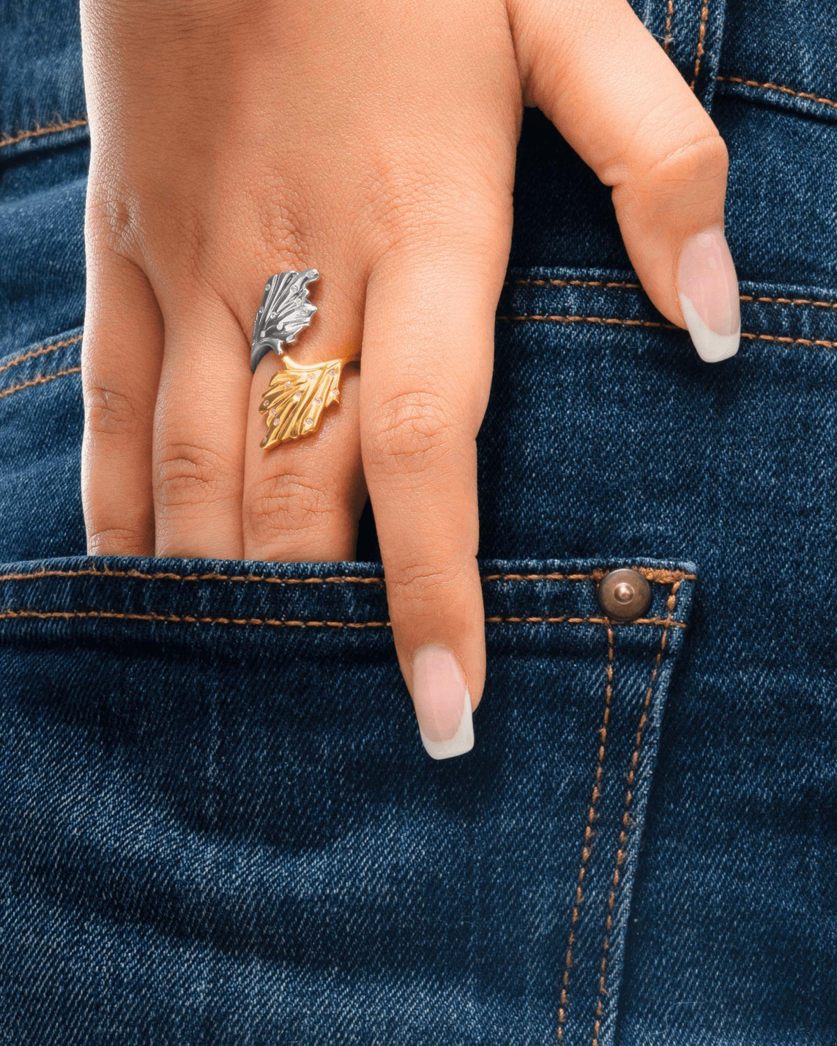 A close-up of a person's hand resting in the pocket of dark blue denim jeans, wearing leaf-shaped ring, inspired by corals in gold and silver tones.