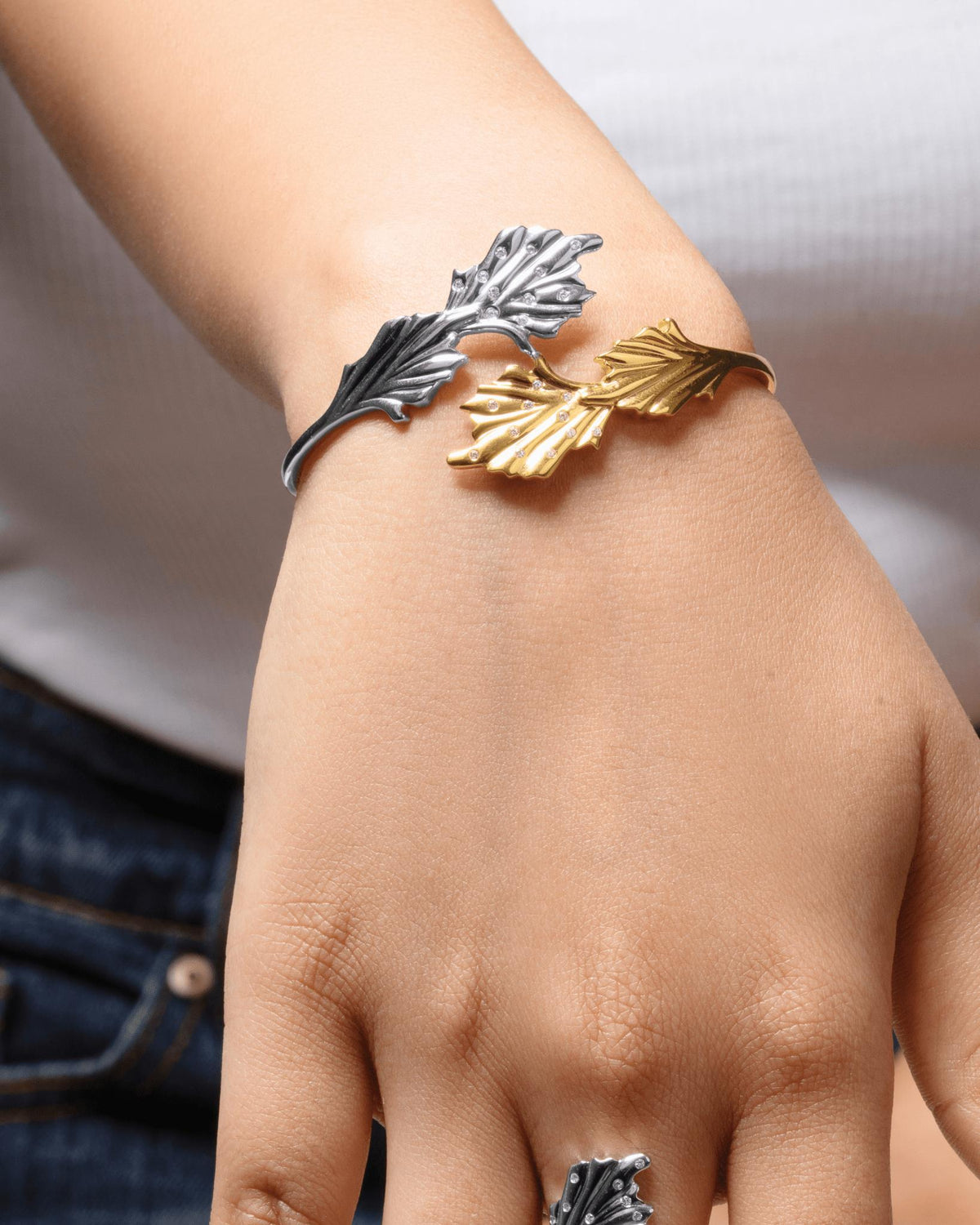 A close-up of a person's wrist wearing a cuff bracelet with three leaf-shaped metal elements in silver, gold, and two-tone silver and gold finishes.