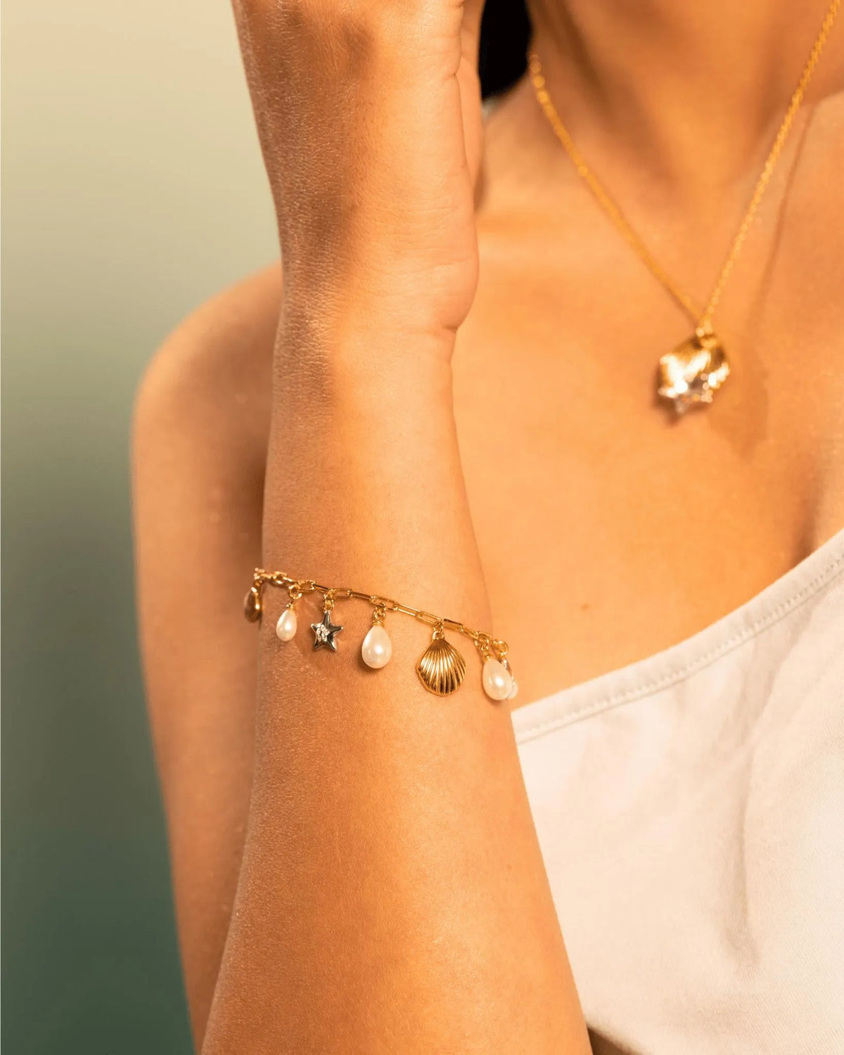 A close-up of a woman's wrist wearing a delicate gold charm bracelet with seashell and star charms, paired with a gold necklace.