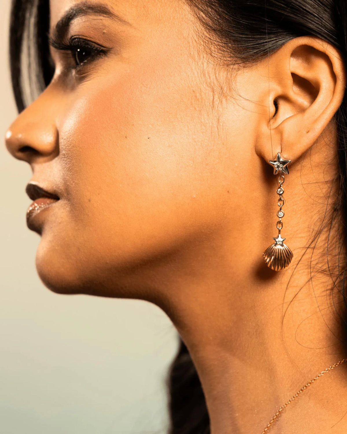A close-up side profile of a woman wearing a dangling earring with a star-shaped top, small clear stones, and a gold shell-shaped drop.