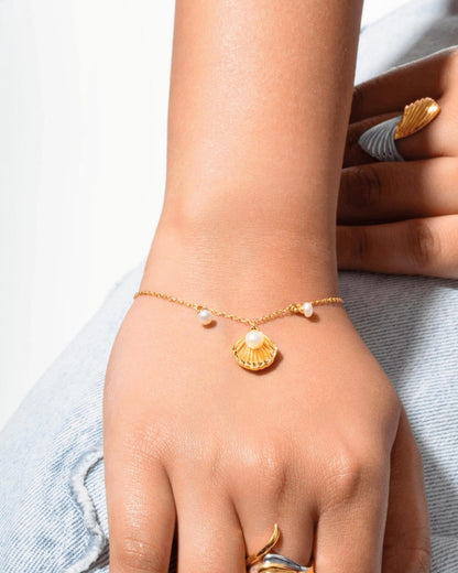 A delicate gold chain bracelet with a scallop shell pendant and small pearl-like beads, shown worn on a person's wrist. The person is also wearing gold rings, and the background includes light blue denim fabric.
