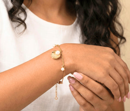 A close-up of a person's wrist wearing a delicate gold chain bracelet with a scallop shell charm, a small white pearl, and a drop pearl, paired with a white top.