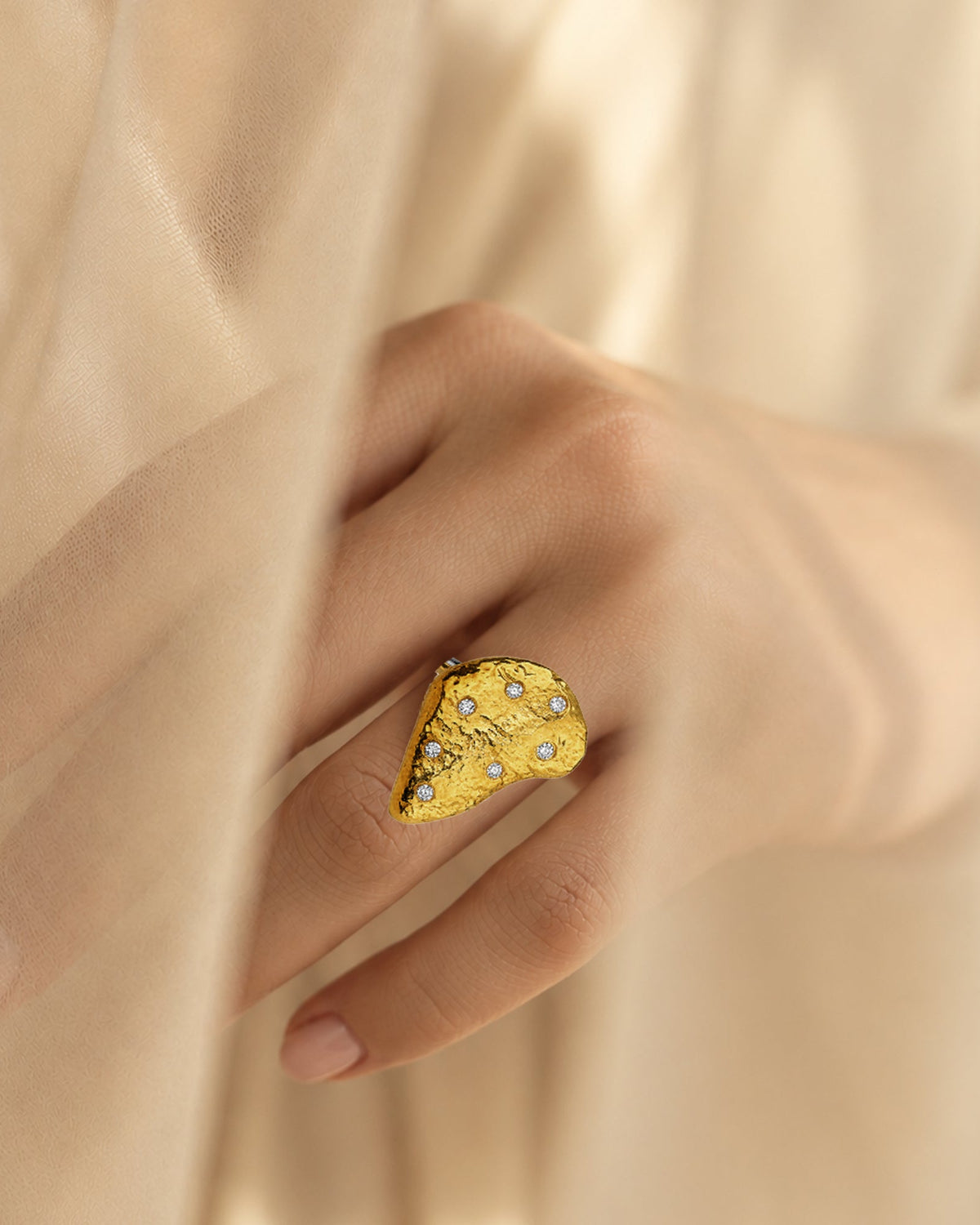 A close-up of a hand wearing a chunky, organic-shaped gold vermeil ring embellished with CZ, soft beige curtain