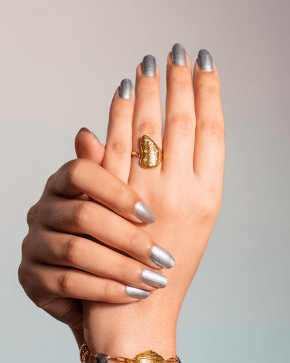 A close-up of a person's hands with silver manicured nails, wearing a textured gold ring, inspired by Oyster's Shell, on the middle finger, set against a neutral light gray background.